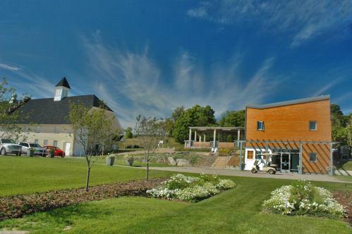 A modern building with wooden accents beside a traditional structure, surrounded by green grass and blooming flowers under a blue sky.