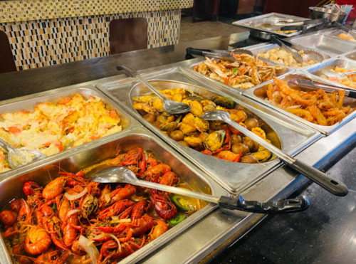 A buffet display featuring various seafood dishes, including crawfish, shrimp, and mixed shellfish in metal trays.