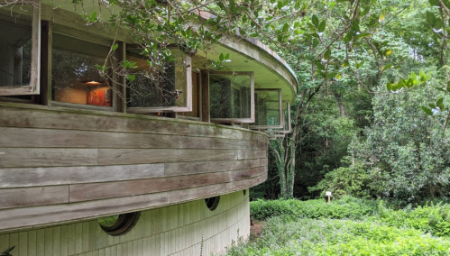 A circular wooden building surrounded by lush greenery and trees, with open windows inviting nature inside.