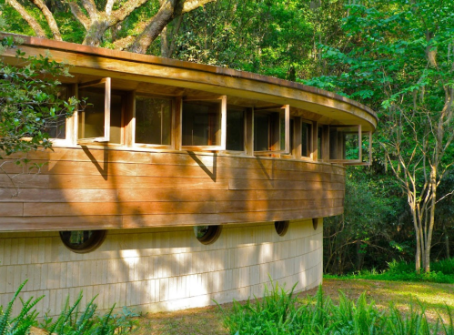 A curved wooden building surrounded by lush greenery and trees, featuring large windows and a unique architectural design.