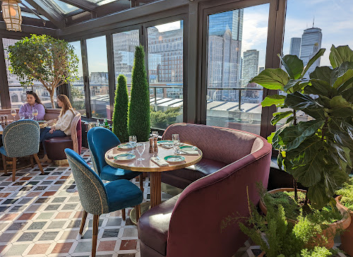 A stylish restaurant interior with a view of the city, featuring plants, elegant seating, and a bright atmosphere.