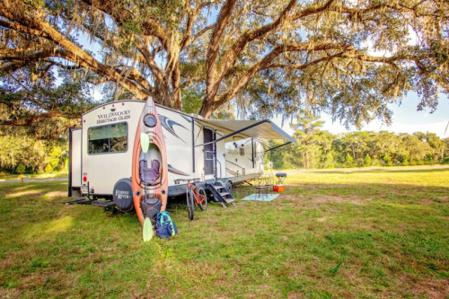 A camper trailer parked under a large tree, with a kayak and outdoor gear nearby on a grassy field.