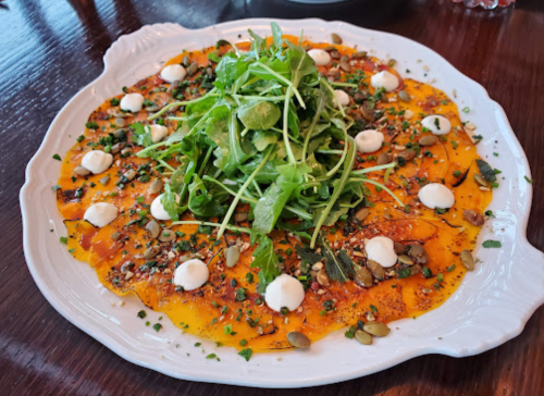 A vibrant dish featuring thinly sliced squash topped with arugula, seeds, herbs, and dollops of creamy sauce.