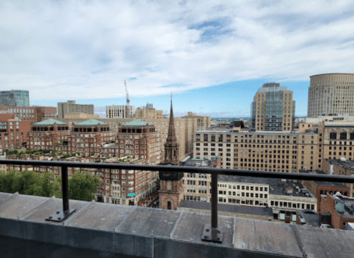 View from a rooftop overlooking a city skyline with buildings and a church spire under a partly cloudy sky.