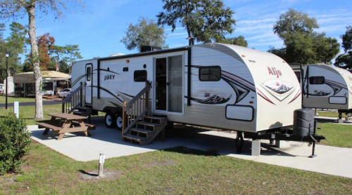 A parked travel trailer with steps, surrounded by grass and trees, in a sunny outdoor setting.