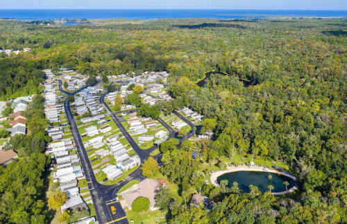 Aerial view of a mobile home community surrounded by lush greenery and a small pond, with the ocean in the distance.