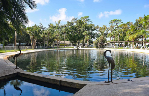 A serene pond surrounded by trees, with two elegant sculptures of cranes at the water's edge under a clear blue sky.