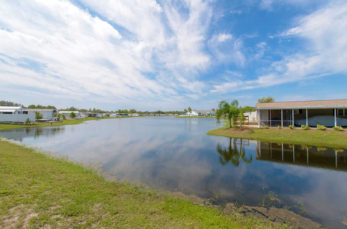 A serene landscape featuring a calm lake, blue sky, and reflections, with houses along the shore.