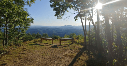 Scenic mountain view with two wooden benches under sunlight, surrounded by trees and rolling hills in the distance.