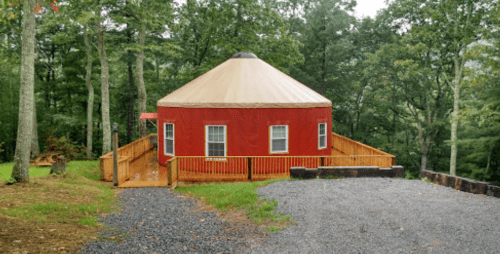 A red yurt with a tan roof, surrounded by trees and a wooden deck, set on a gravel path.