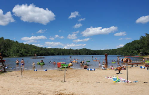 A sunny beach scene with people swimming, lounging, and enjoying the water at a lake surrounded by trees.