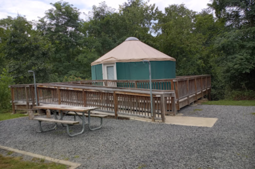 A wooden deck surrounds a green yurt with a tan roof, set in a wooded area with a picnic table nearby.