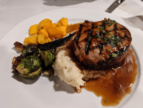 A plated meal featuring grilled pork chop, mashed potatoes, Brussels sprouts, and diced mango.