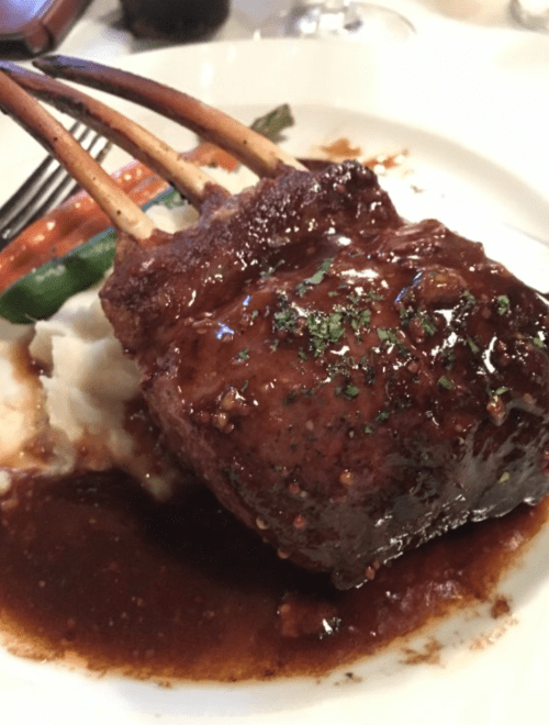 A beautifully plated rack of lamb with a rich sauce, served on a bed of mashed potatoes and garnished with vegetables.