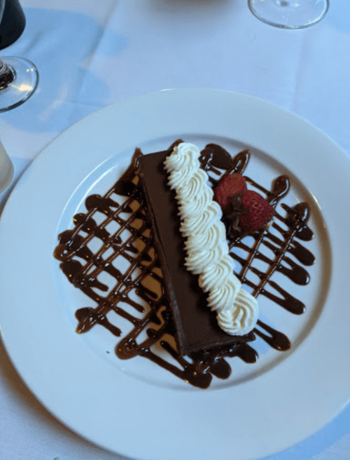 A chocolate dessert on a white plate, topped with whipped cream and garnished with strawberries, drizzled with chocolate sauce.
