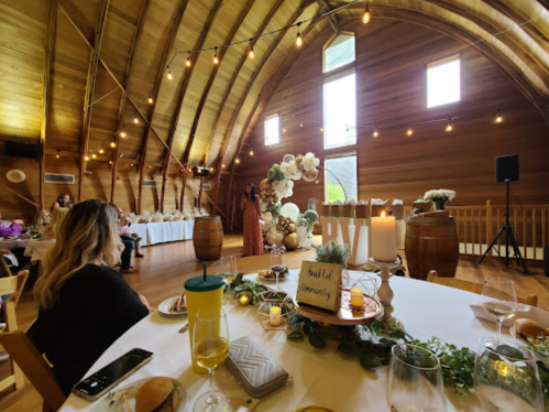 A woman speaks at a rustic venue decorated with flowers and lights, while guests sit at tables with drinks.