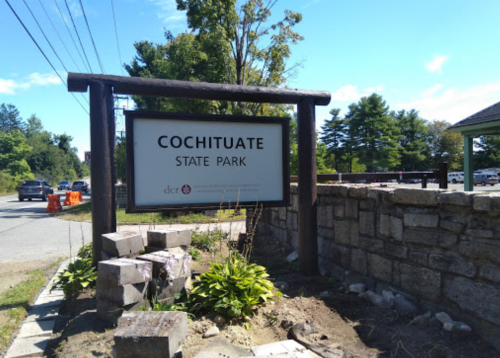 Sign for Cochituate State Park, surrounded by greenery and stone, with a road and vehicles in the background.