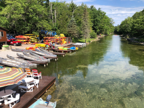 A scenic view of a calm river lined with colorful kayaks and canoes, surrounded by lush greenery and a sunny sky.