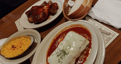 A plate with lasagna topped with cheese, a side of macaroni and cheese, and BBQ chicken wings with bread rolls.