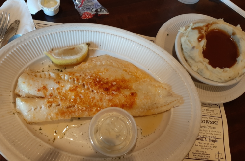 A plate with two fillets of fish topped with seasoning, a lemon wedge, and a side of mashed potatoes with gravy.