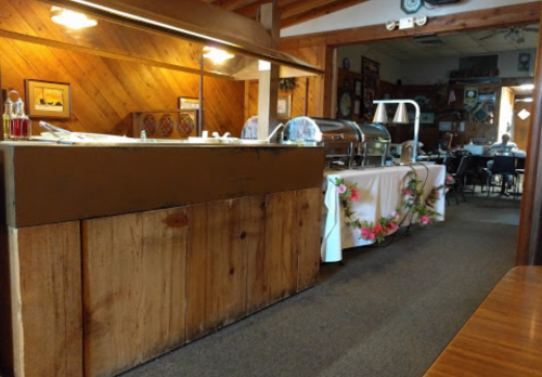 A rustic dining area with a buffet setup, wooden walls, and tables in the background.