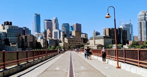 A sunny day view of a city skyline with people walking along a bridge, surrounded by buildings and greenery.
