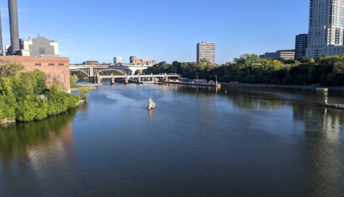 A calm river flows past a city skyline, with bridges and greenery lining the banks under a clear blue sky.