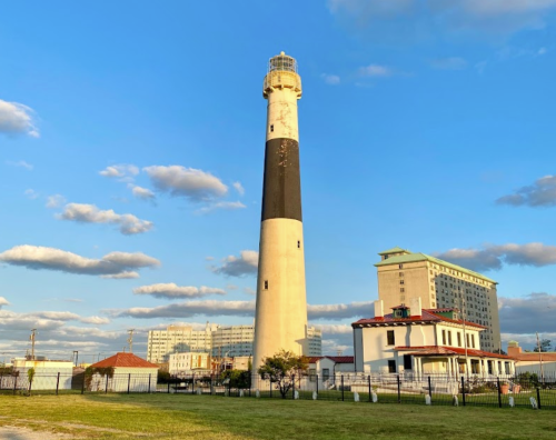 A tall lighthouse with a black and white striped tower stands beside a building under a blue sky with scattered clouds.