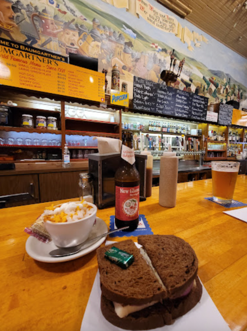 A sandwich on a plate with a side of popcorn, a beer, and a bottle on a wooden bar with a mural in the background.