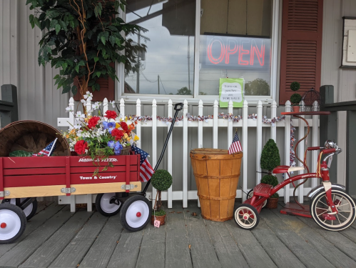 A welcoming porch display with a red wagon, flowers, a wooden barrel, and a vintage tricycle, all under an "OPEN" sign.