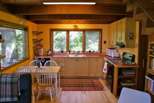 Cozy kitchen with wooden cabinets, a dining table, and large windows overlooking greenery. Warm, inviting atmosphere.