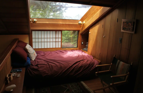 Cozy wooden bedroom with a large window, a bed with maroon bedding, and a chair in a serene, natural setting.