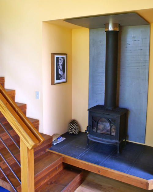 A cozy corner with a black wood stove, framed art, and a staircase made of wood, set against a yellow wall.