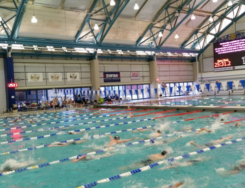 A busy indoor swimming pool with swimmers in lanes, flags overhead, and a scoreboard displaying times.