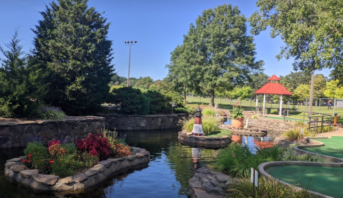 A serene park scene featuring a pond, colorful flowers, and a gazebo under a clear blue sky.