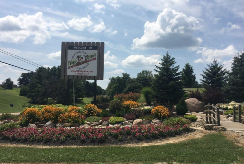 A welcoming sign for a venue surrounded by colorful flowers and greenery under a partly cloudy sky.