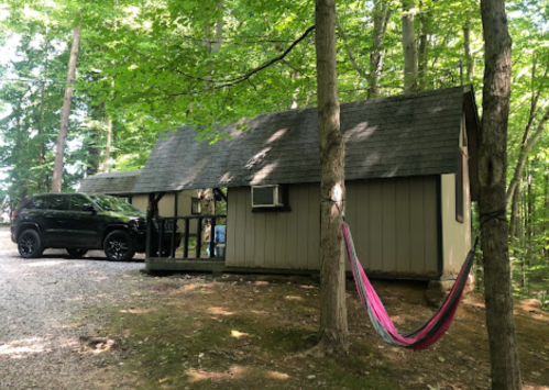 A cozy cabin nestled in a wooded area, with a hammock hanging between trees and a black SUV parked nearby.