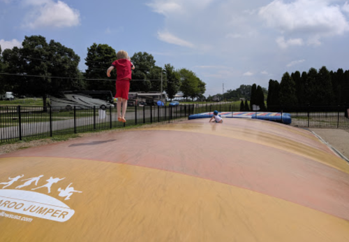 A child in a red outfit jumps on a large inflatable bounce pad, with trees and a road in the background.