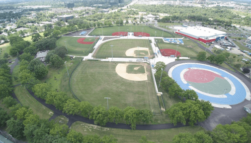 Aerial view of a sports complex featuring baseball fields, a track, and green spaces surrounded by trees.