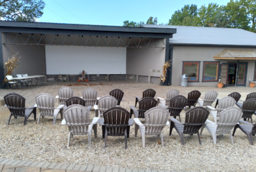 Outdoor seating area with rows of chairs facing a blank screen, surrounded by gravel and greenery.