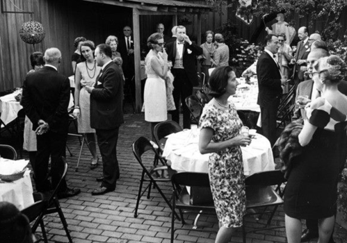 A black-and-white photo of a lively outdoor gathering with guests mingling around tables in a garden setting.