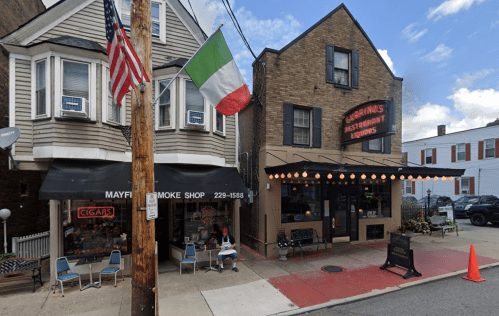A street view featuring a smoke shop and an Italian restaurant with outdoor seating and decorative lights.