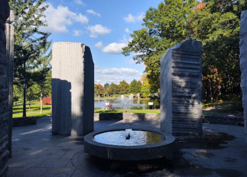 A serene park scene featuring stone sculptures, a circular fountain, and a pond surrounded by trees and blue skies.