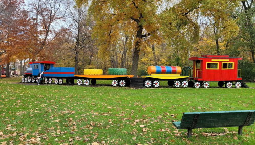 Colorful train play structure in a park, surrounded by autumn leaves and trees. A green bench is visible in the foreground.