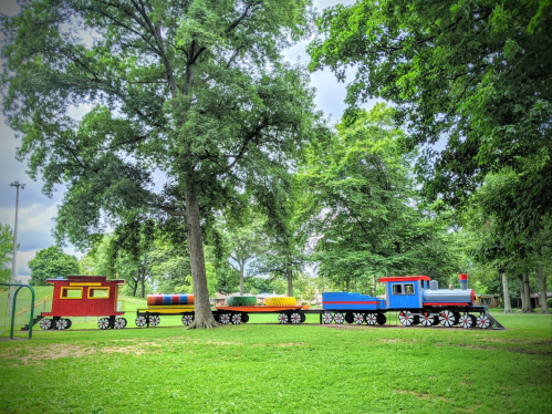 Colorful train play structure in a park, surrounded by green grass and large trees under a cloudy sky.