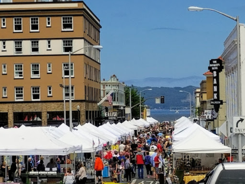 A bustling street market with white tents, crowds of people, and buildings lining the street, set against a scenic backdrop.