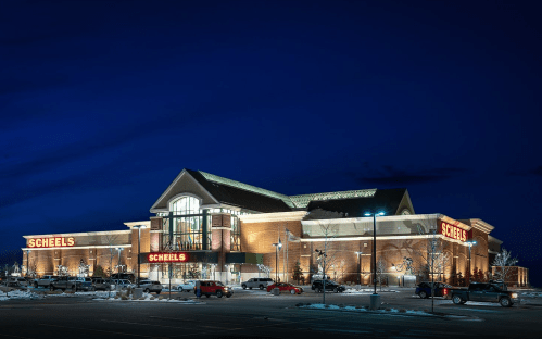 A large Scheels store illuminated at night, with parked cars and a clear sky in the background.