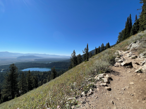 A scenic view from a mountain trail, featuring a lake and lush greenery under a clear blue sky.