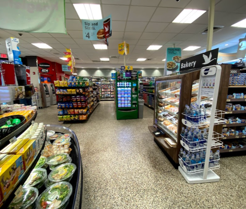A well-stocked convenience store aisle featuring snacks, drinks, and a bakery display. Brightly lit and organized layout.