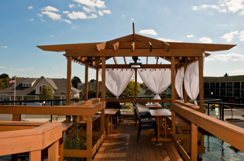 A wooden gazebo with white curtains, overlooking a scenic view under a blue sky with scattered clouds.